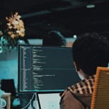 A person sits at a desk working on a computer with code displayed on the monitor in a dimly lit office environment.