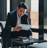 A man in a suit sits on a chair, looking at a tablet. There is a coffee cup and a notebook on the table in front of him.