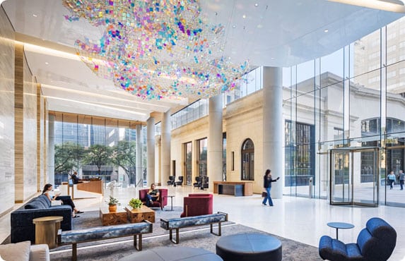 Spacious, modern building lobby with large windows, seating areas, a reception desk, and a colorful abstract ceiling installation. Several people are present in the space.